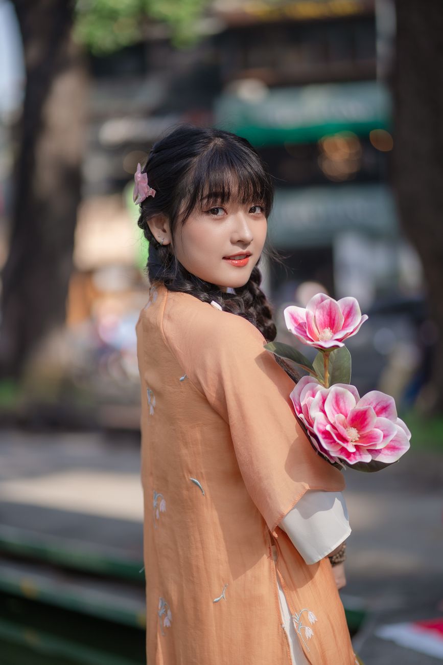 Charming woman wearing orange Ao Dai holding pink flowers outdoors, embodying cultural elegance.