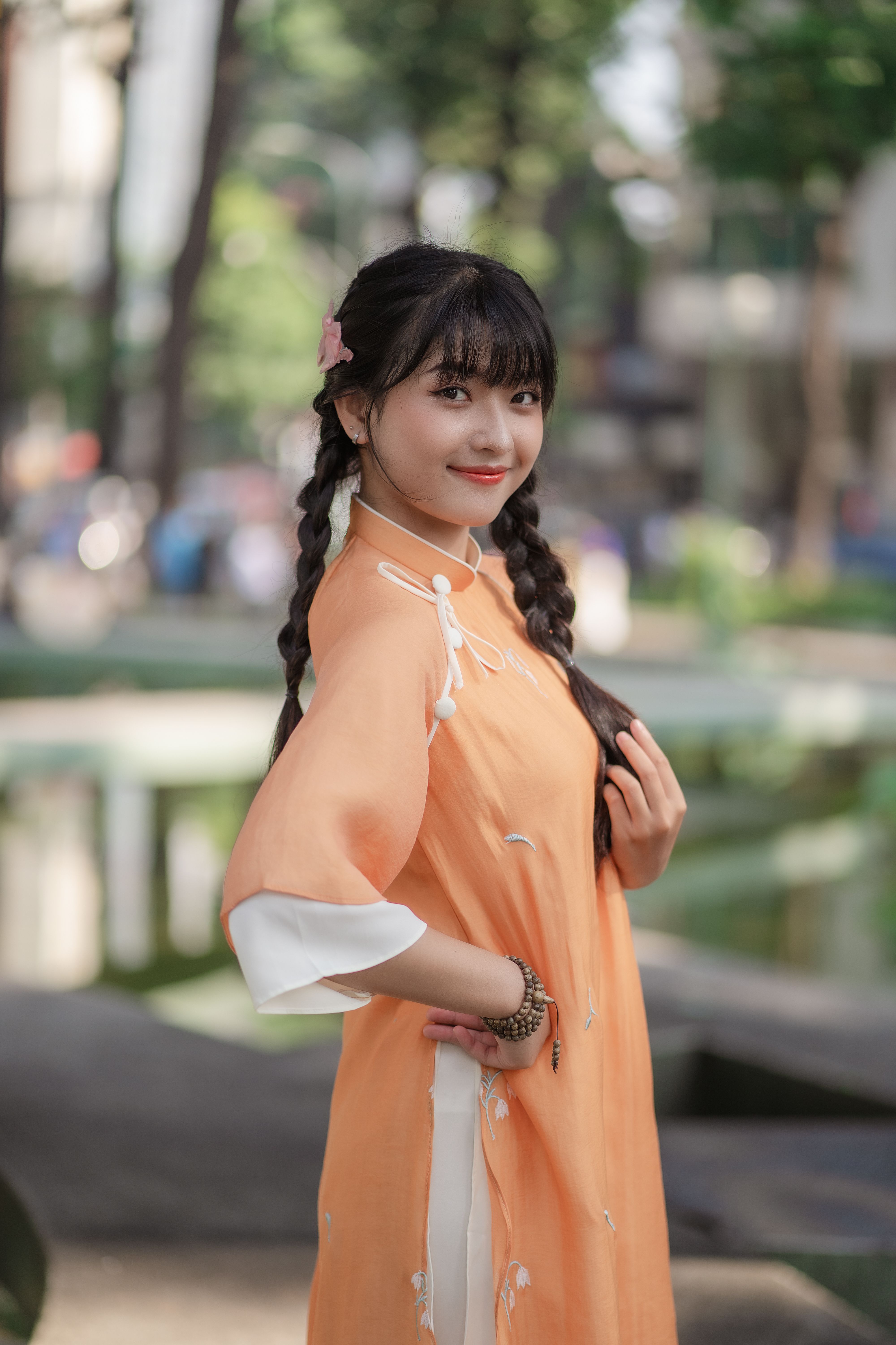 A young woman wearing an orange ao dai poses gracefully in an outdoor park setting.