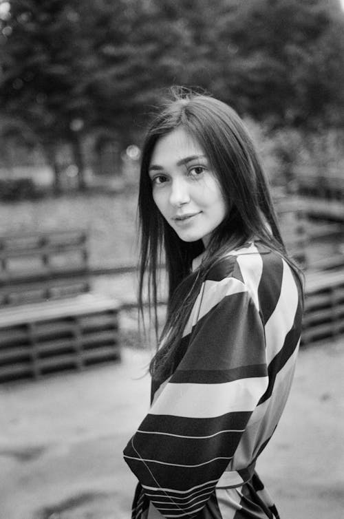 Black and white of casual brunette in striped dress looking kindly at camera standing against benches in park