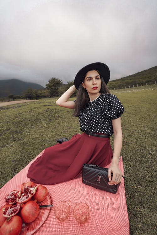 Woman in a Black Polka-dot Blouse and Burgundy Skirt Sitting on a Picnic Blanket in the Mountains