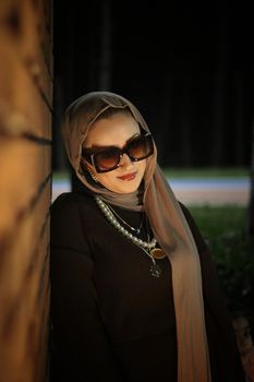 Chic portrait of a stylish woman in hijab and sunglasses leaning against a wall at night.