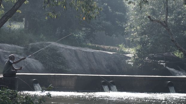 An elderly man enjoys fishing by a tranquil river amidst lush greenery.
