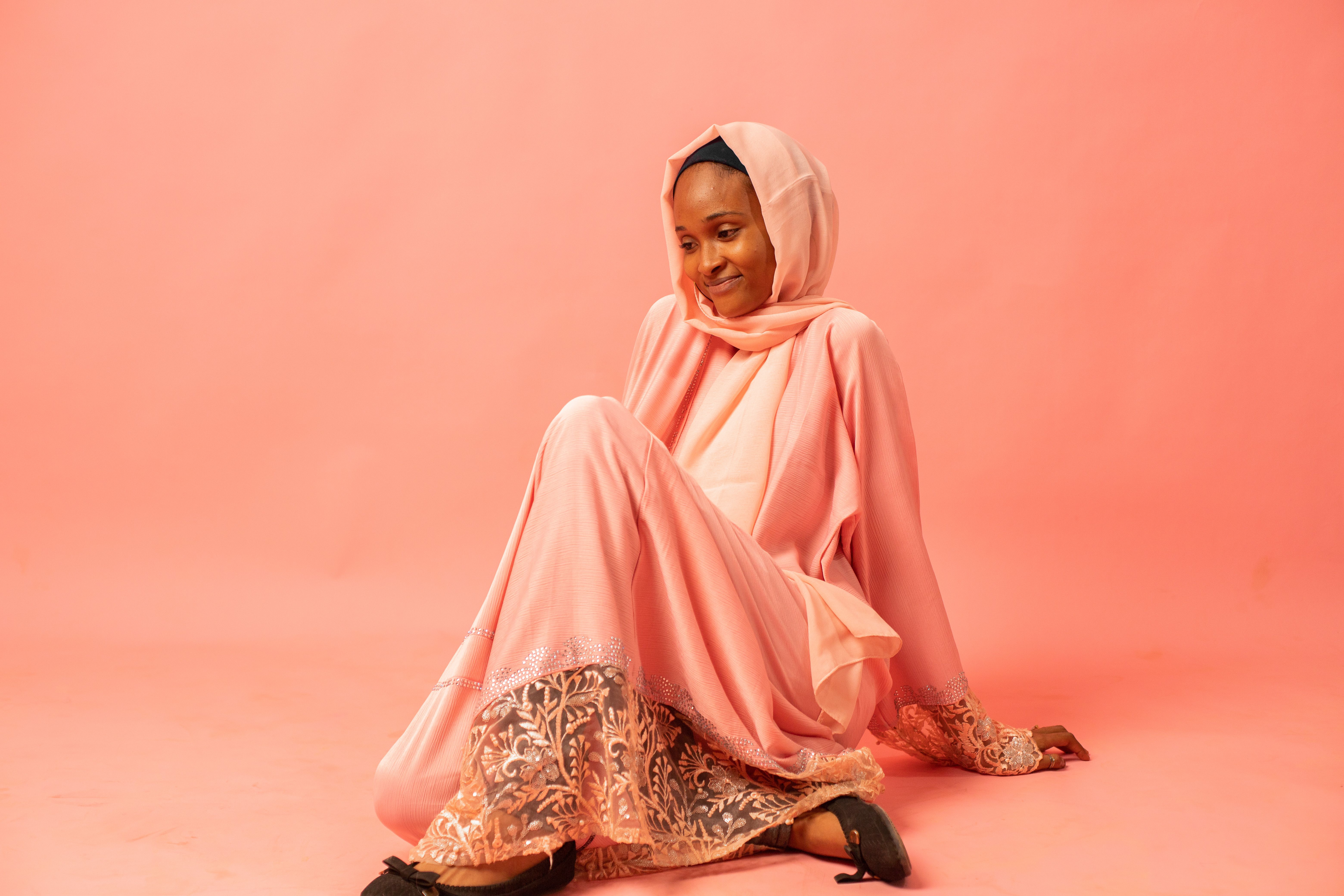A woman in a pink traditional dress with a headscarf, sitting in a studio setting.