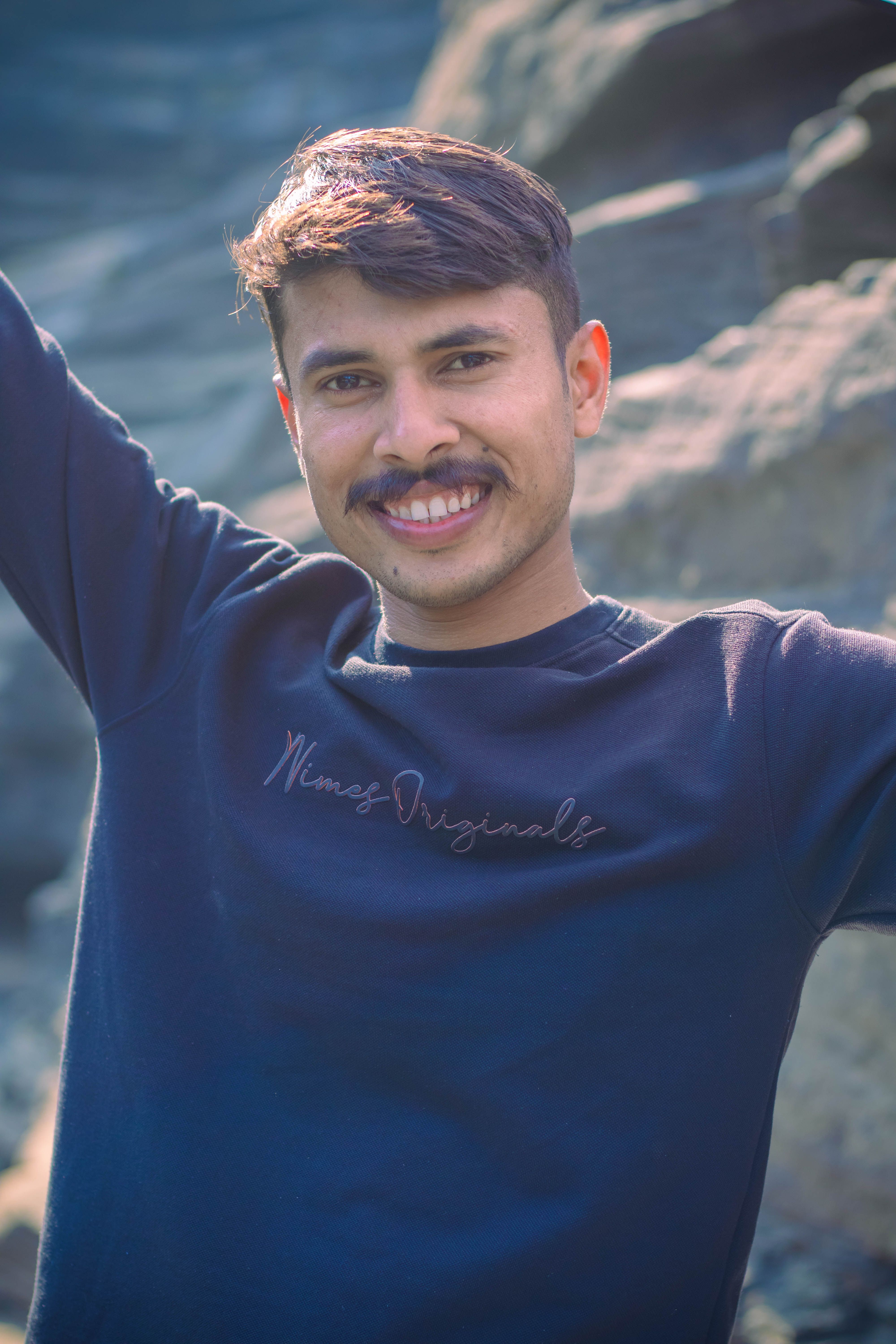Young man with a mustache smiling and posing outdoors in casual attire.