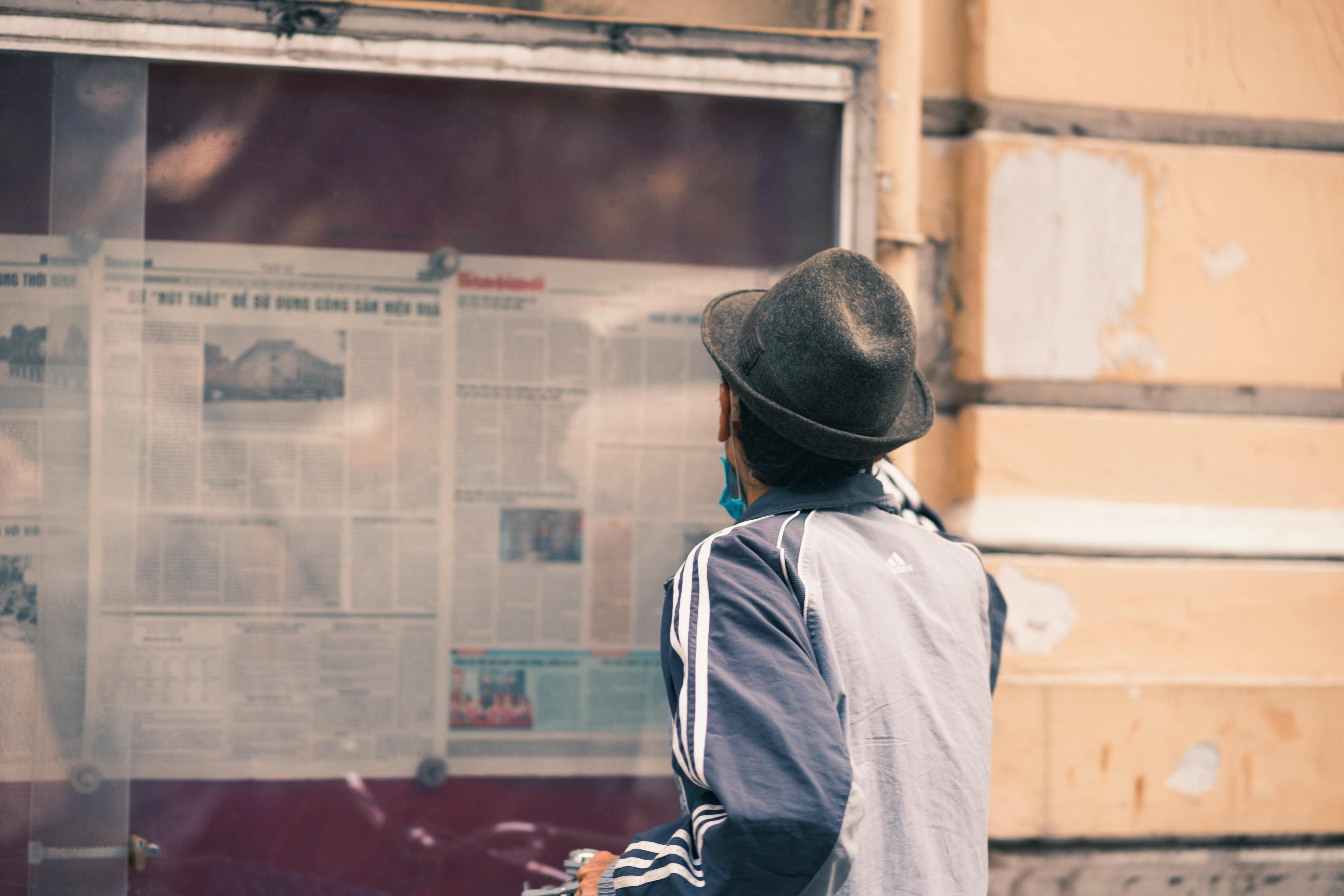 back of a man in a hat reading a newspaper pinned to a board