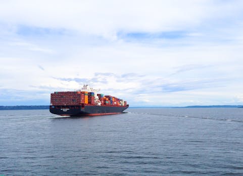 A vast container ship loaded with colorful cargo sails across a calm ocean under a cloudy sky.