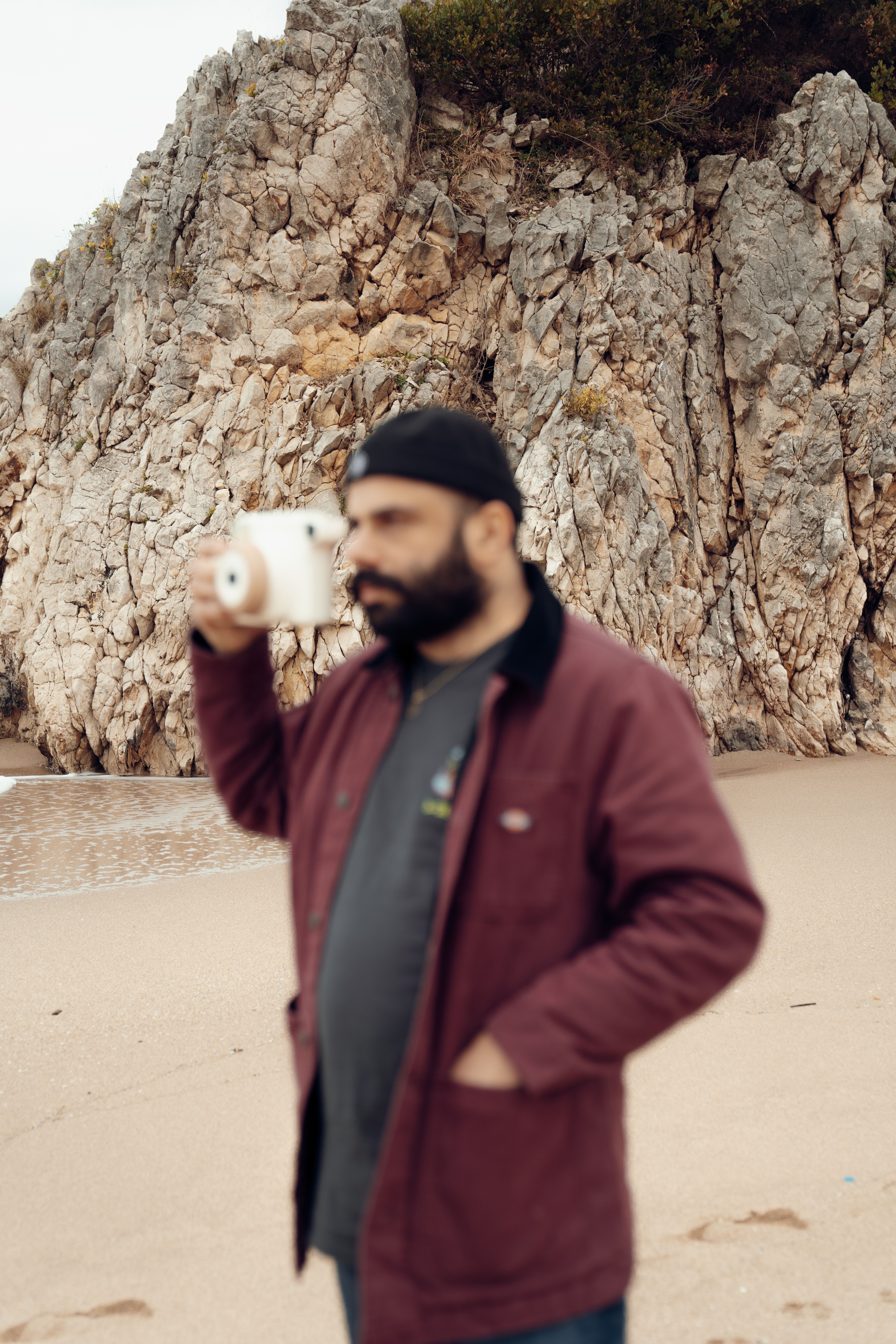 A man holding a camera stands defocused on a rocky beach, creating a soft and artistic composition.