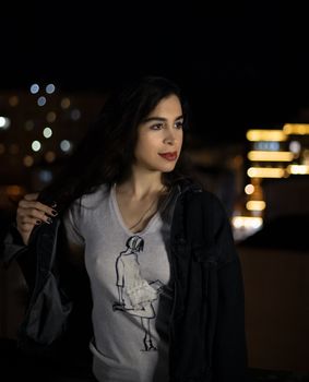 Stylish young woman in a denim jacket and graphic tee posing in an urban night setting.