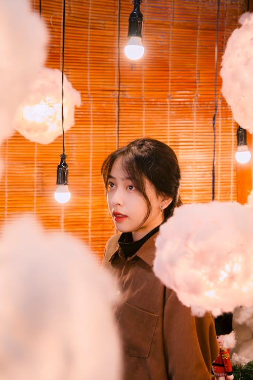 Brunette Woman in Brown Jacket Standing Among Hanging Light Bulbs