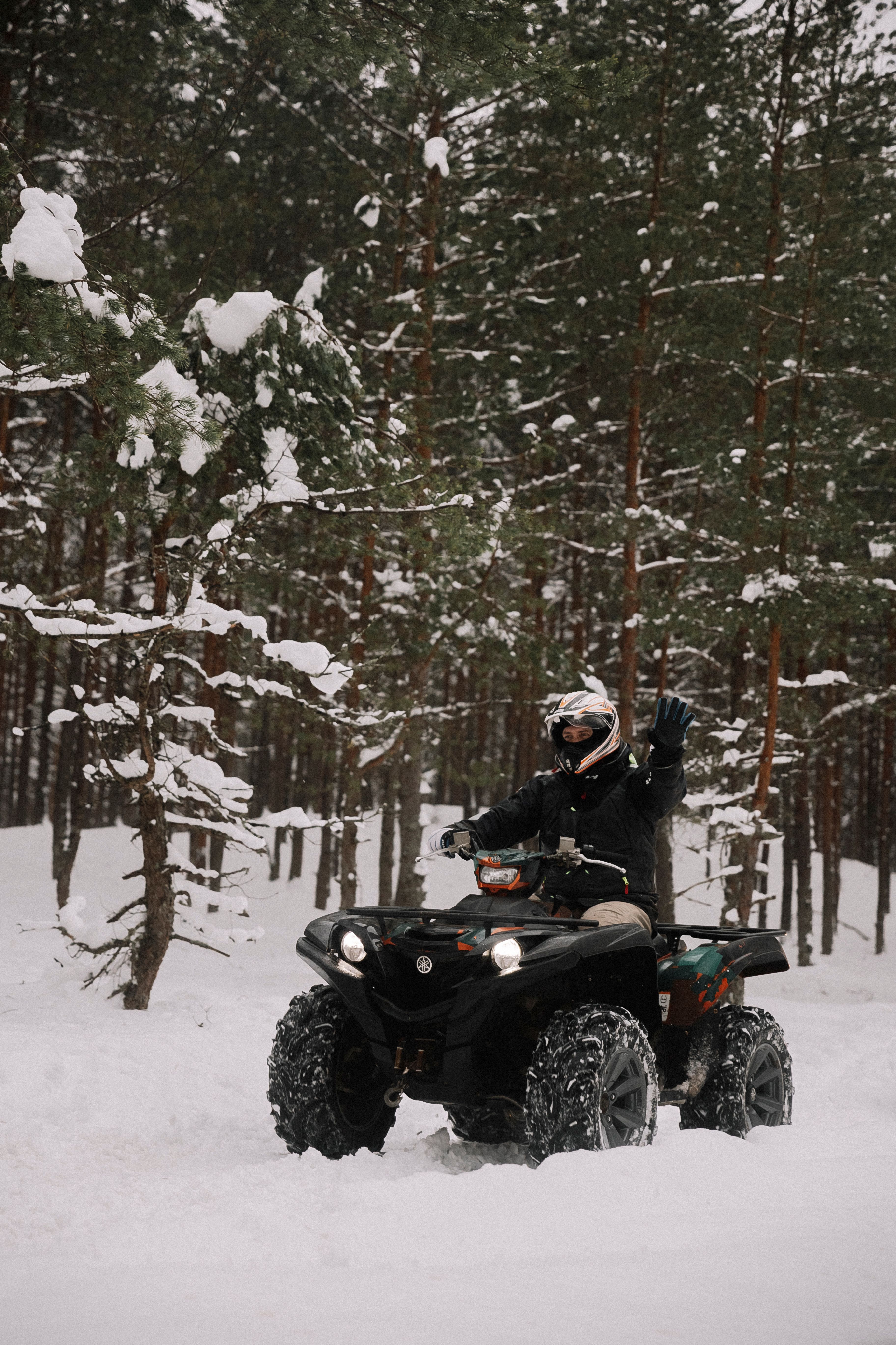 Man riding an ATV in a snow-covered forest, waving with excitement in a winter landscape.