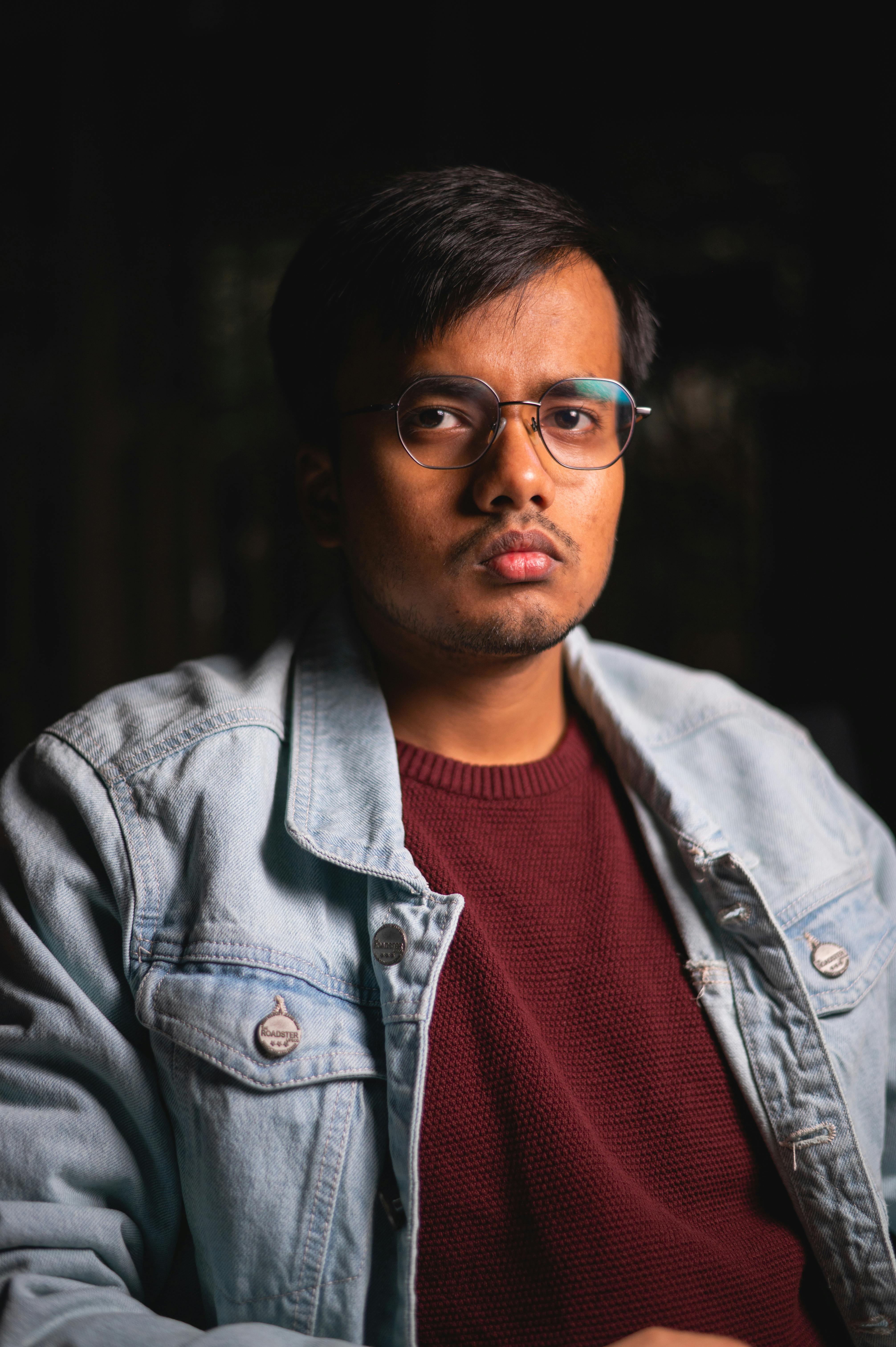 Dramatic portrait of a young man wearing glasses and denim jacket against a dark background.