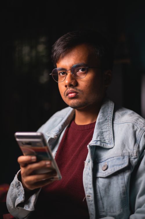 Free Portrait of a young man with glasses holding a smartphone, wearing a jean shirt indoors. Stock Photo
