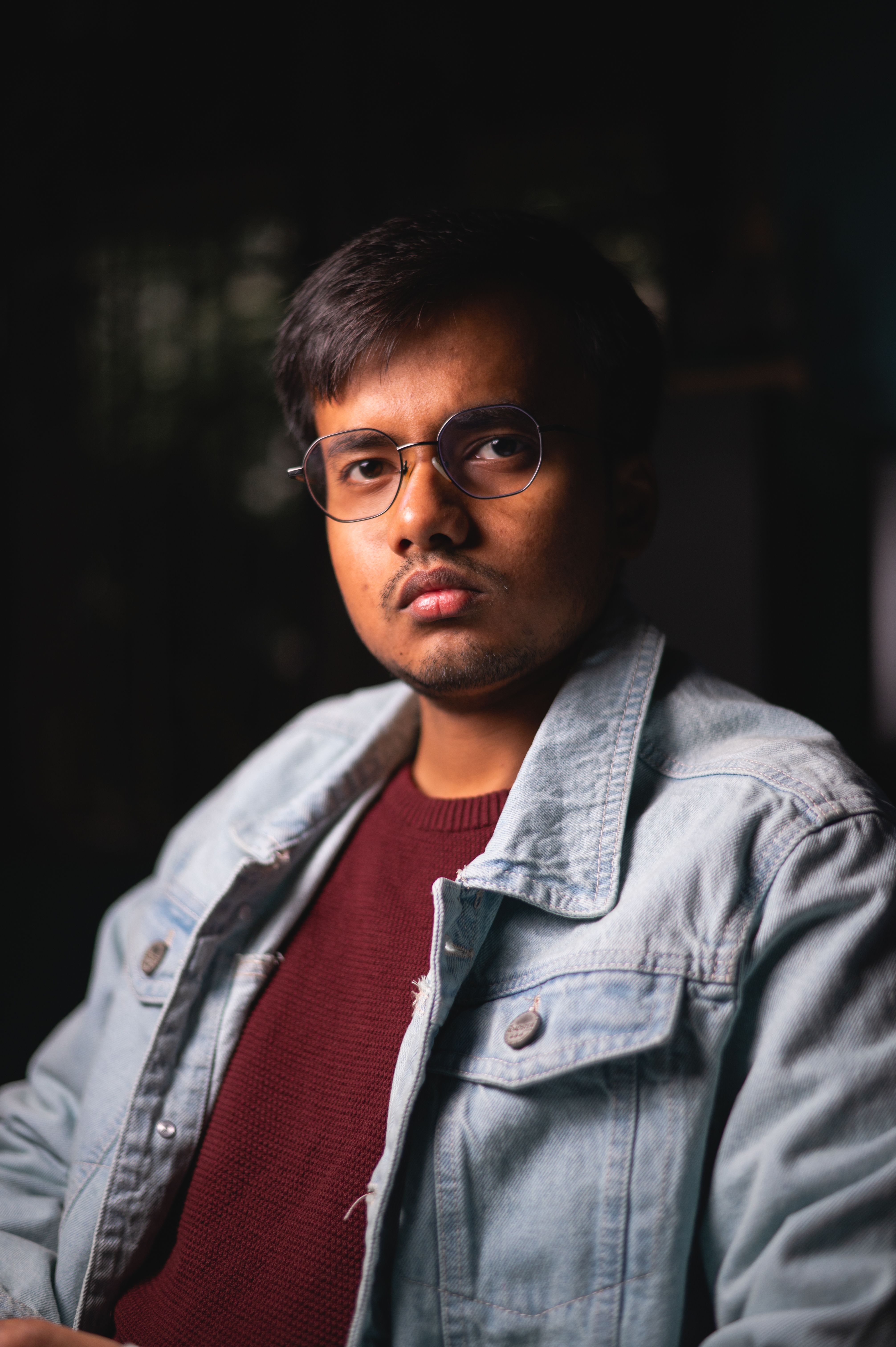 A young man with eyeglasses and a mustache wearing a denim jacket looks serious in a moody portrait.