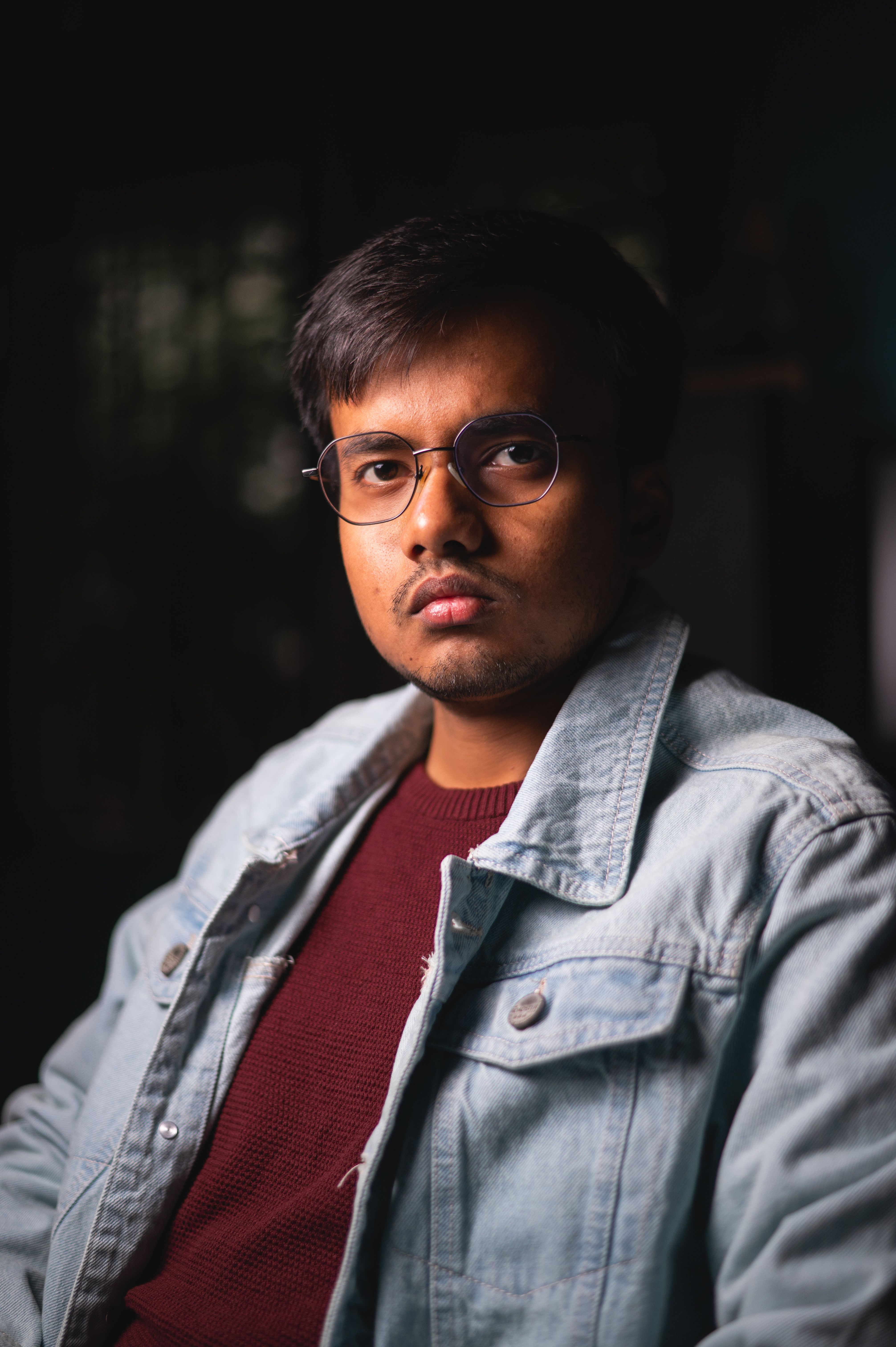 Portrait of a young man in a jean jacket and glasses set against a dark background.