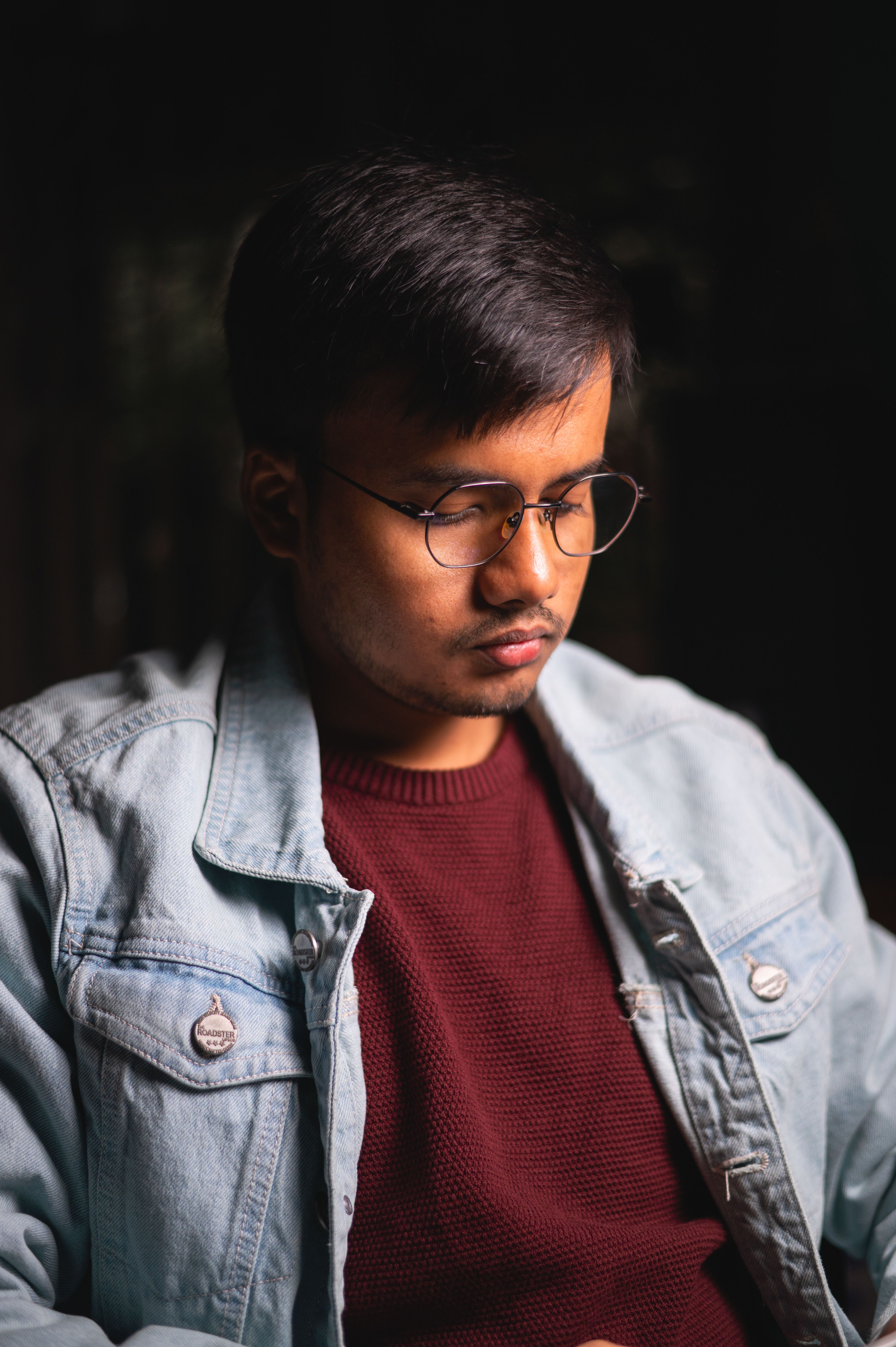 Portrait of a serious young man wearing a denim jacket and glasses, looking contemplative indoors.