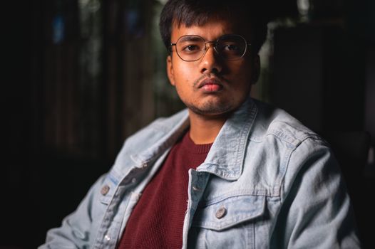 Portrait of a serious young man wearing a denim jacket and eyeglasses indoors.