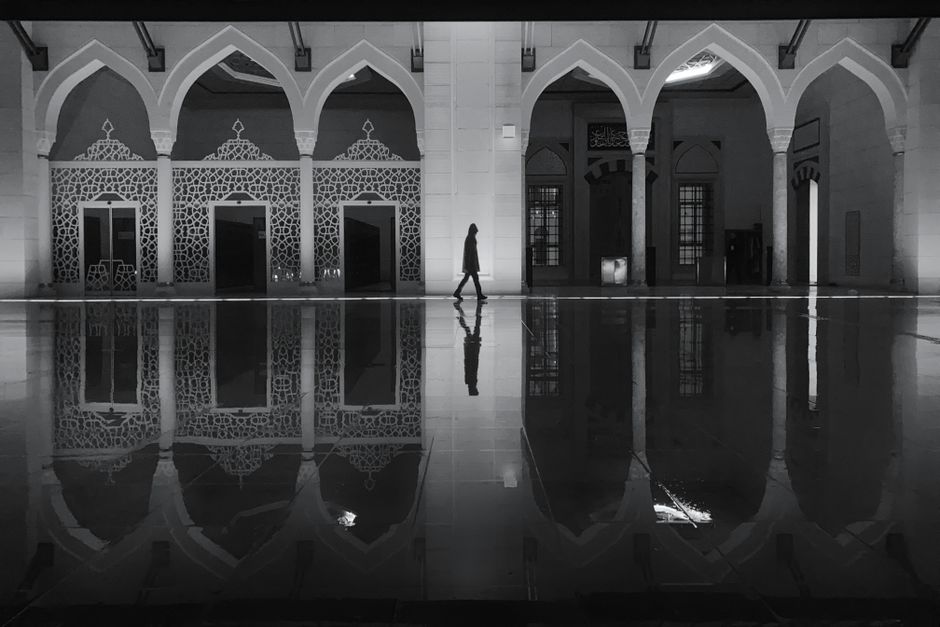 Silhouette of a person walking in a mosque corridor with decorative arches and reflections on the floor at night.