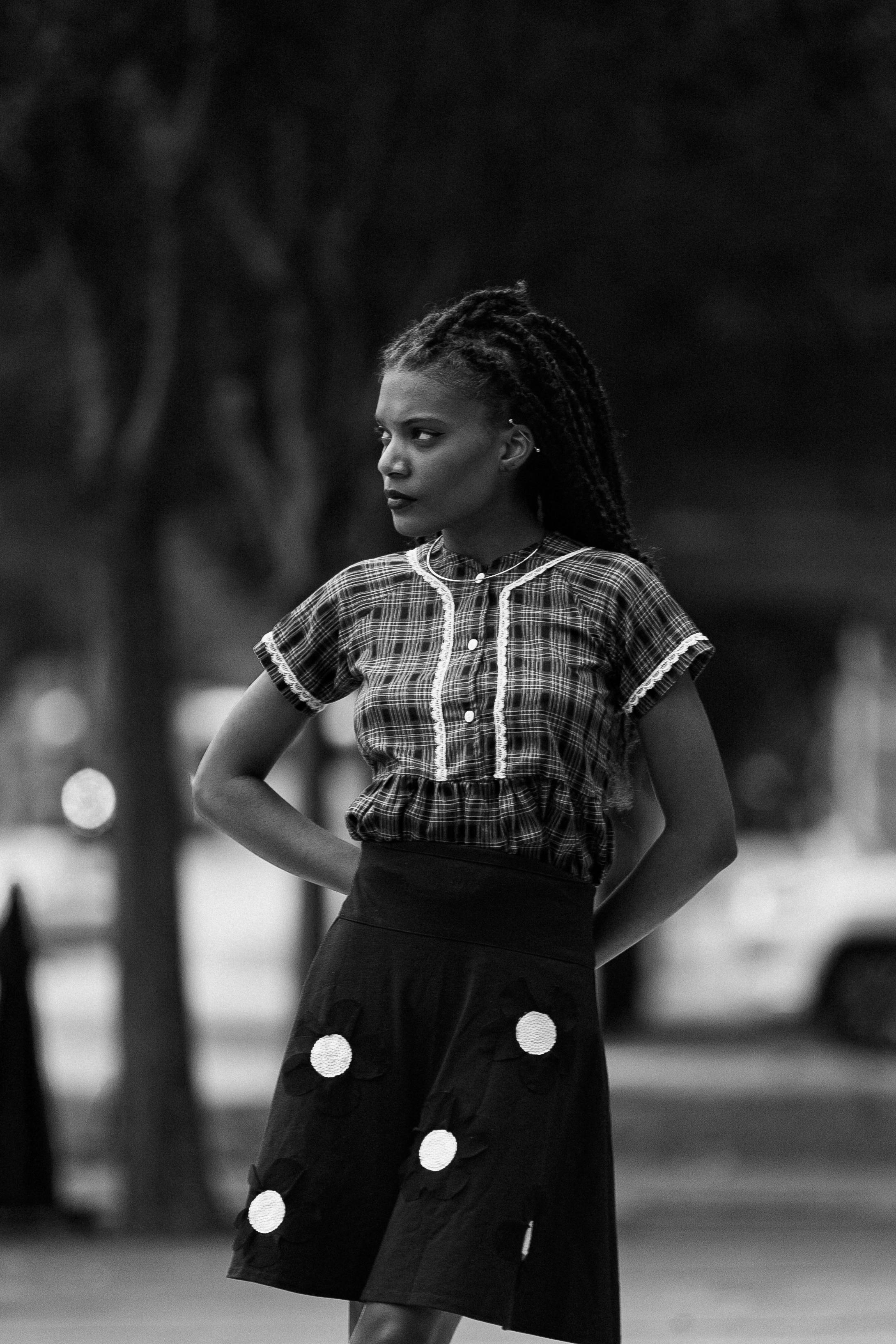 black and white photo of a young woman in a checkered shirt and dotted skirt standing outside