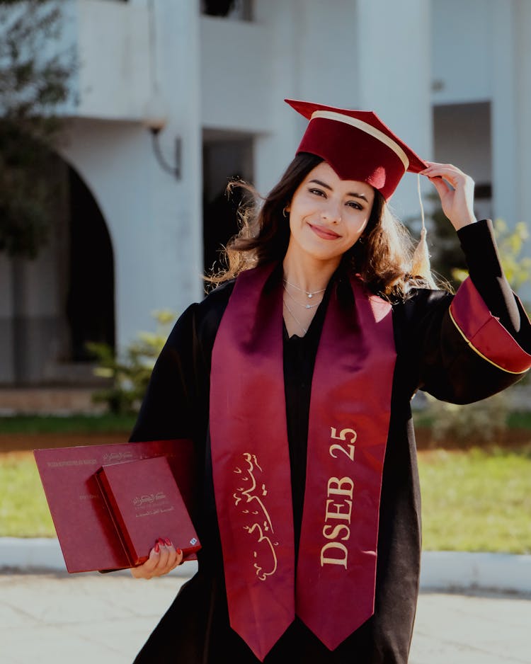 Young Woman In A Graduation Gown And Mortarboard Standing Outside 