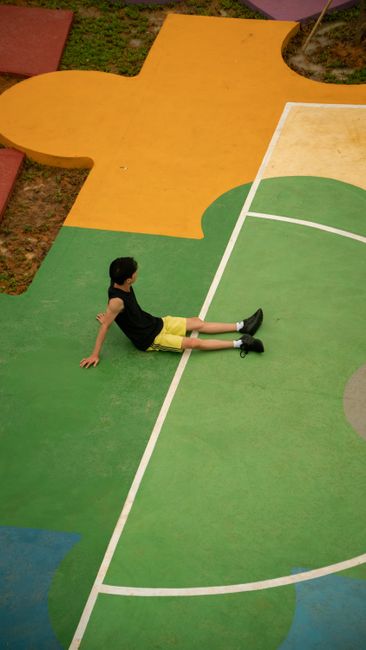 A man in a tank top sits on a vibrant puzzle-shaped playground court. Aerial view.