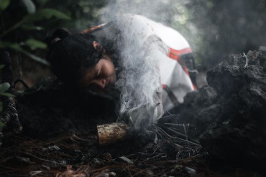 A woman with black hair tends to a small smoky fire in a forest setting, creating a serene mood in Guatemala.