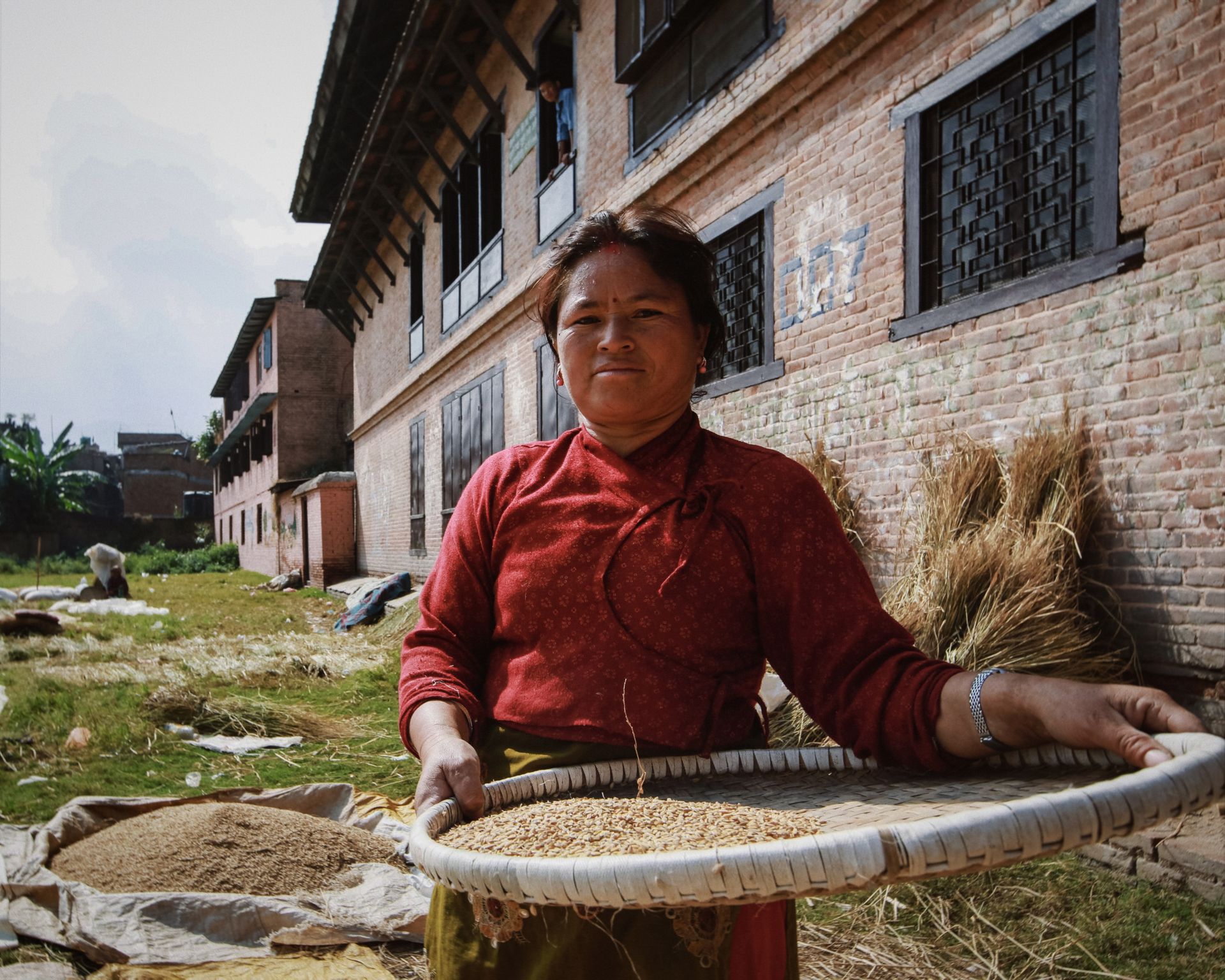 A woman in traditional attire sorts grain using a sieve outdoors near a rustic building.