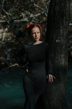 Red-haired woman in a black dress standing by a tree in a forest with sunlight creating a mystical atmosphere.