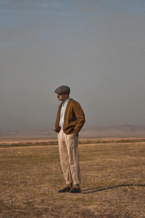 Free A man with classic attire and hat standing in a vast, open meadow on a sunny day. Stock Photo