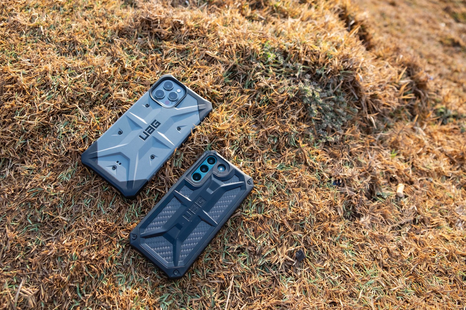 Two rugged UAG smartphone cases placed on a grassy forest floor, showcasing durability.