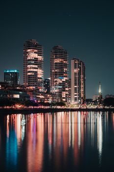 A vibrant urban skyline at night with illuminated skyscrapers reflecting in calm waters.