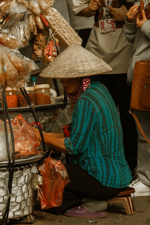Elderly Woman with Chinese Hat Kneeling by Stall at Market