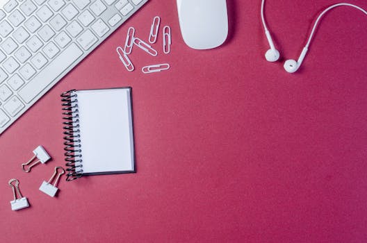 Flat lay of office supplies on a pink background featuring a keyboard, mouse, notepad, and earphones.
