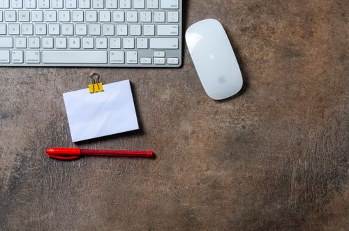 Free Flat lay of a minimalist office desk setup featuring a keyboard, mouse, red pen, and note. Stock Photo