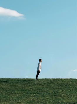 A man stands atop a grassy hill leaning against a clear blue sky. Minimalist, calm scene.