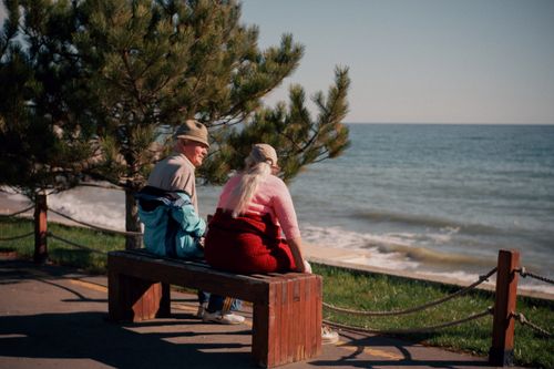 Free Elderly couple sitting on a bench by the coast, enjoying a sunny summer day by the ocean. Stock Photo