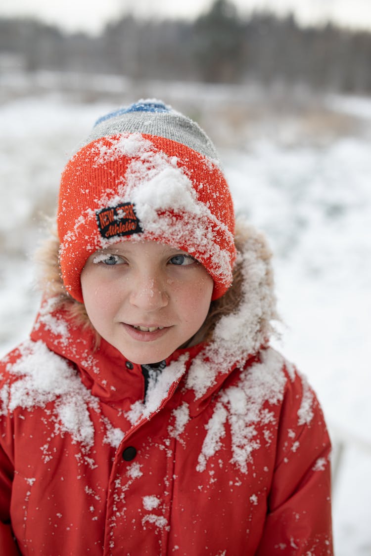 A Boy In A Jacket And Hat Standing On A Snowy Field 