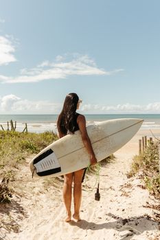 A woman stands with a surfboard facing the ocean on a sunny beach, ready for surfing.