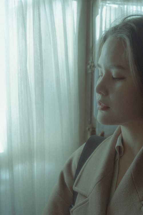 Free Serene woman with closed eyes by a window with soft lighting, creating a peaceful ambiance. Stock Photo