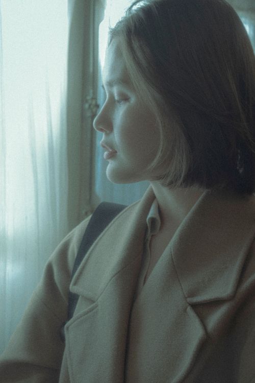 Free A contemplative woman in a coat stands by a window, softly illuminated by natural light. Stock Photo