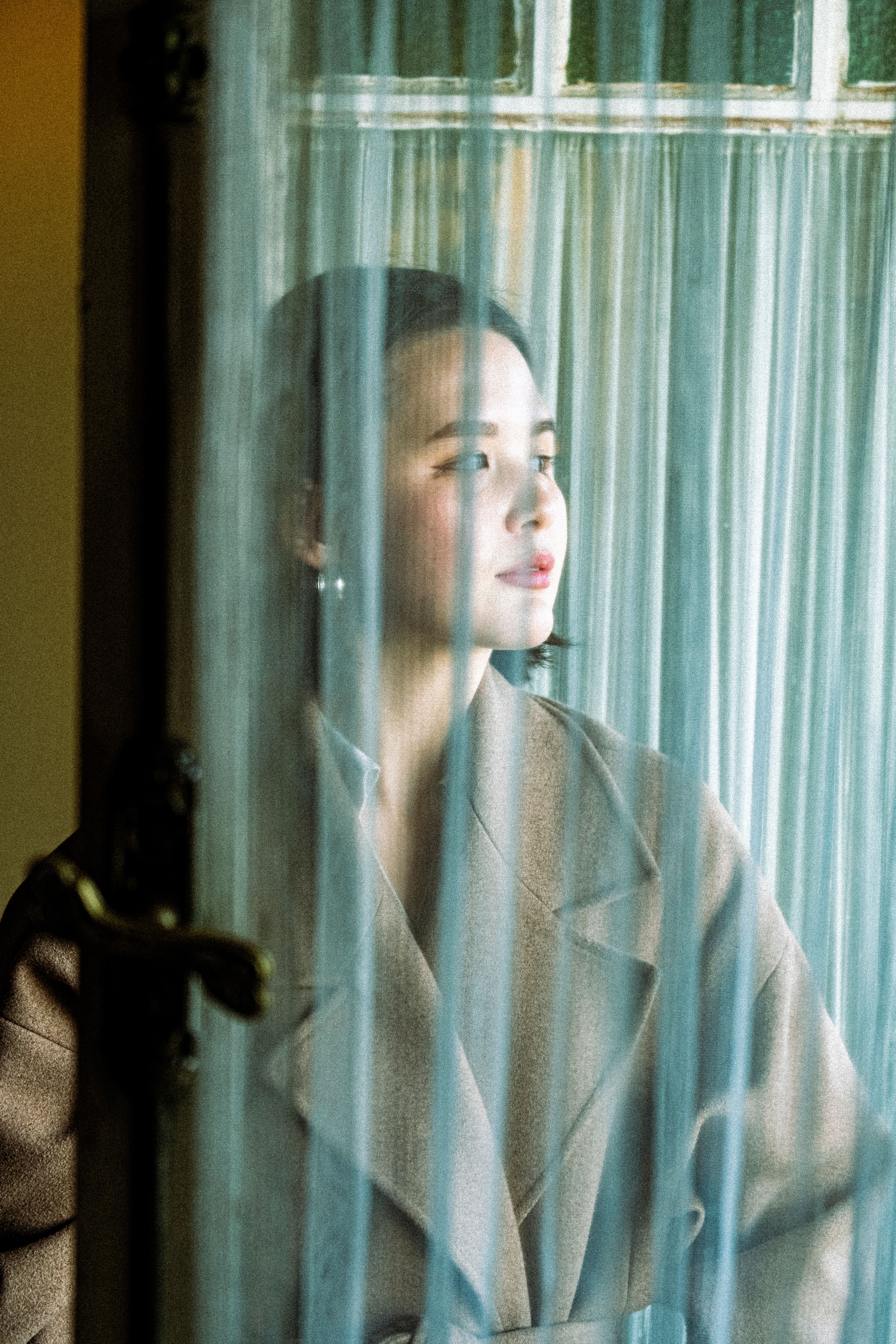 An Asian woman in a coat gazing thoughtfully through sheer curtains near a window.