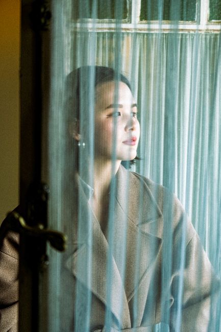 An Asian woman in a coat gazing thoughtfully through sheer curtains near a window.