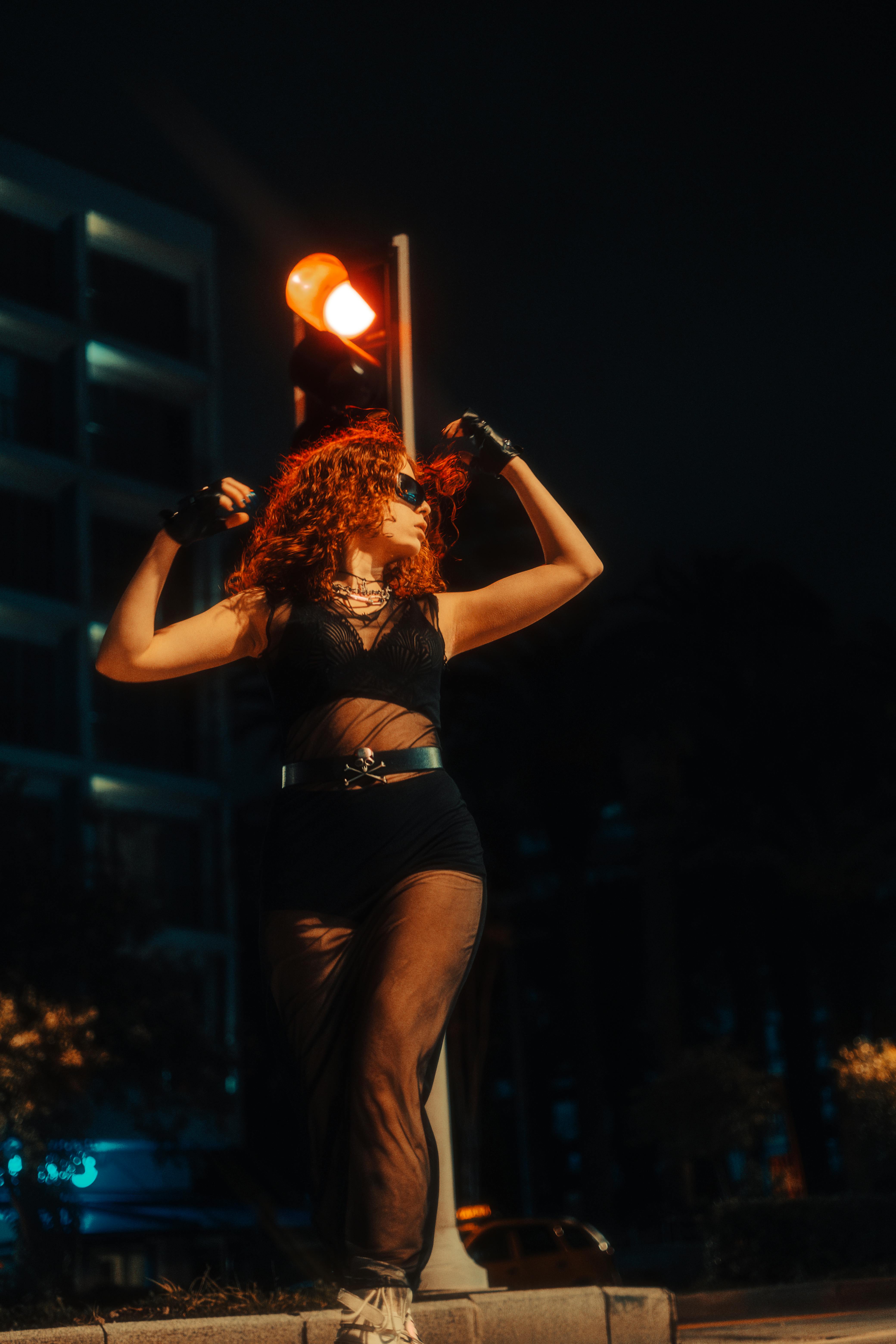 Free Woman posing in sheer outfit under city lights, embodying nightlife fashion. Stock Photo