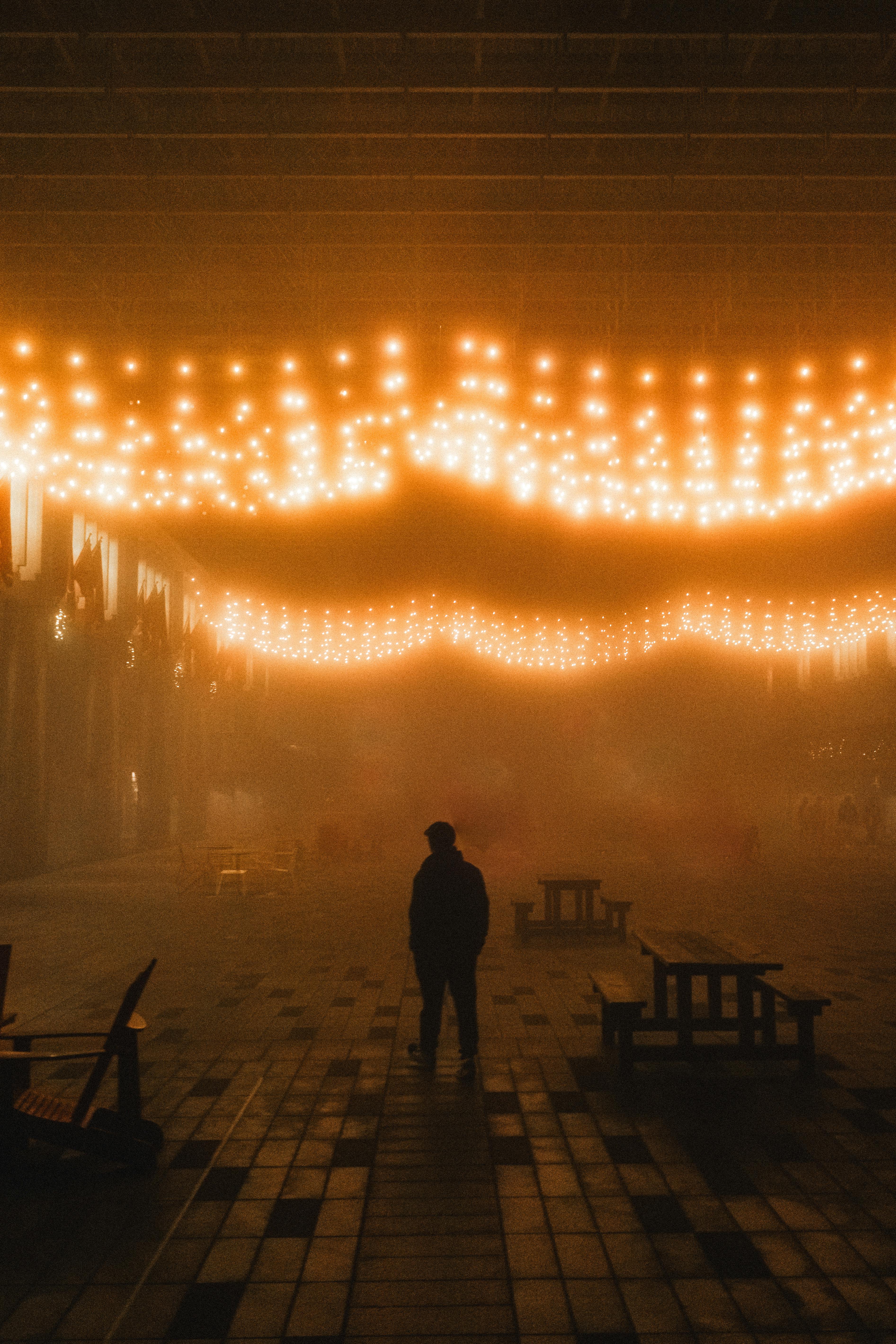 A lone figure stands under warm, glowing string lights in a foggy, atmospheric alley. Perfect mood shot.