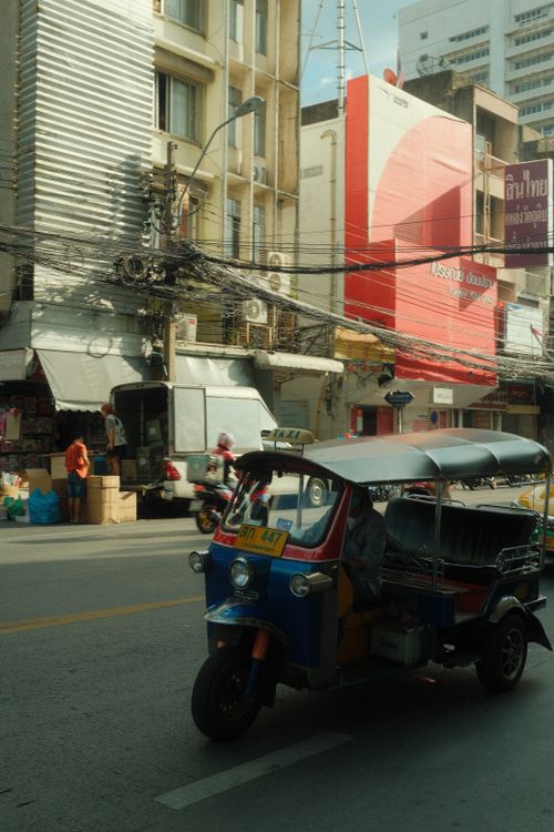 Free Tuk-tuk driving through a busy city street lined with shops and power lines. Stock Photo