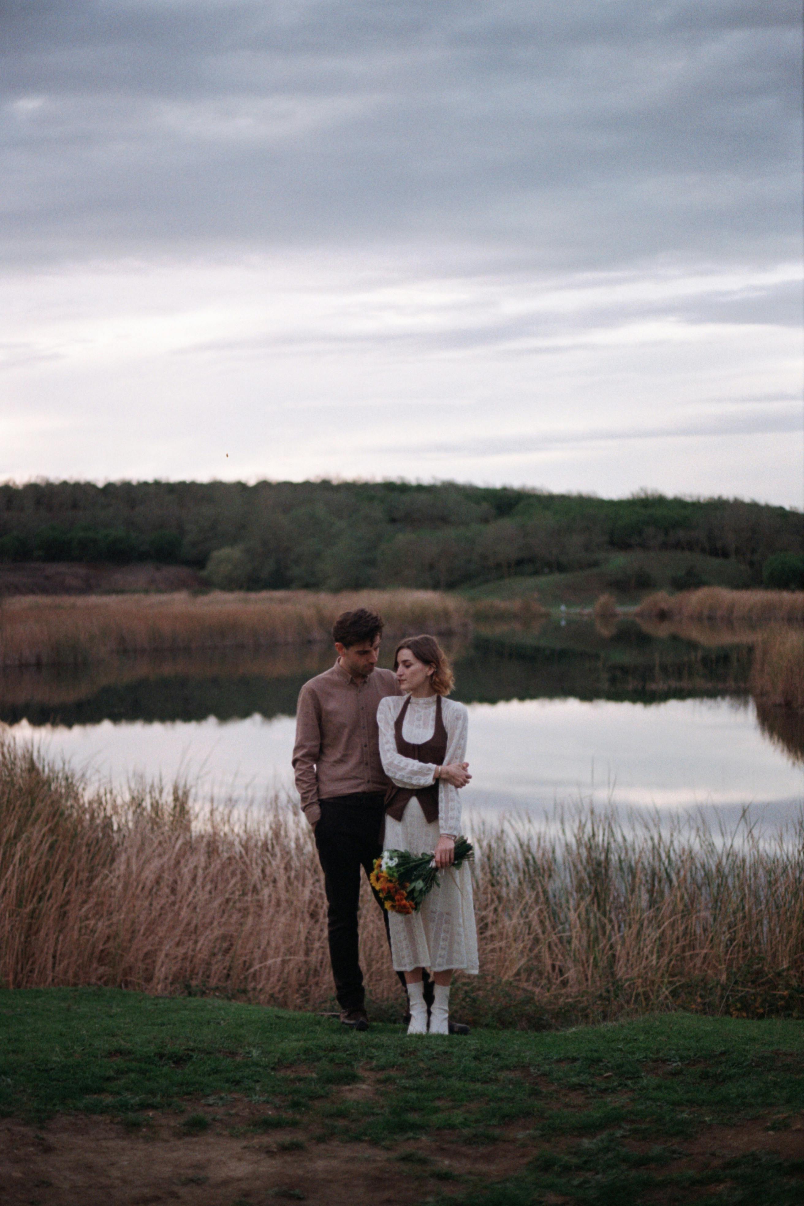 A couple walks hand in hand near a tranquil lake surrounded by reeds, exuding romance and peace.