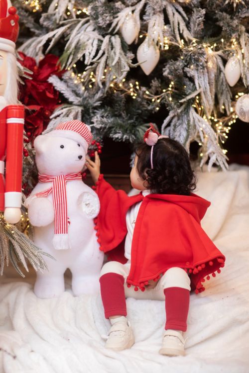 Free Adorable child in red cape sitting by a Christmas tree with festive decorations and a white bear plushie. Stock Photo
