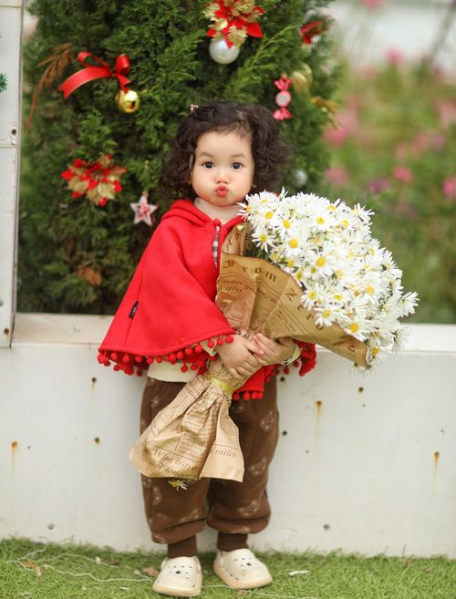 Free Playful child in red coat holds bouquet near decorated Christmas tree outdoors. Stock Photo