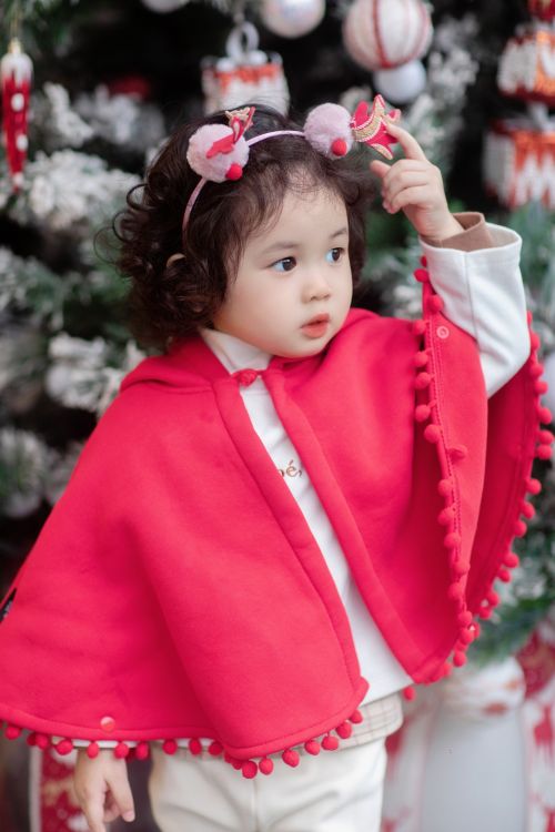 Free Cute child wearing a red poncho with a festive headband in front of a Christmas tree. Stock Photo