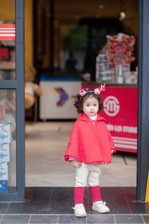 Free A cute toddler girl wearing a red poncho and headband standing in a shop entrance. Stock Photo
