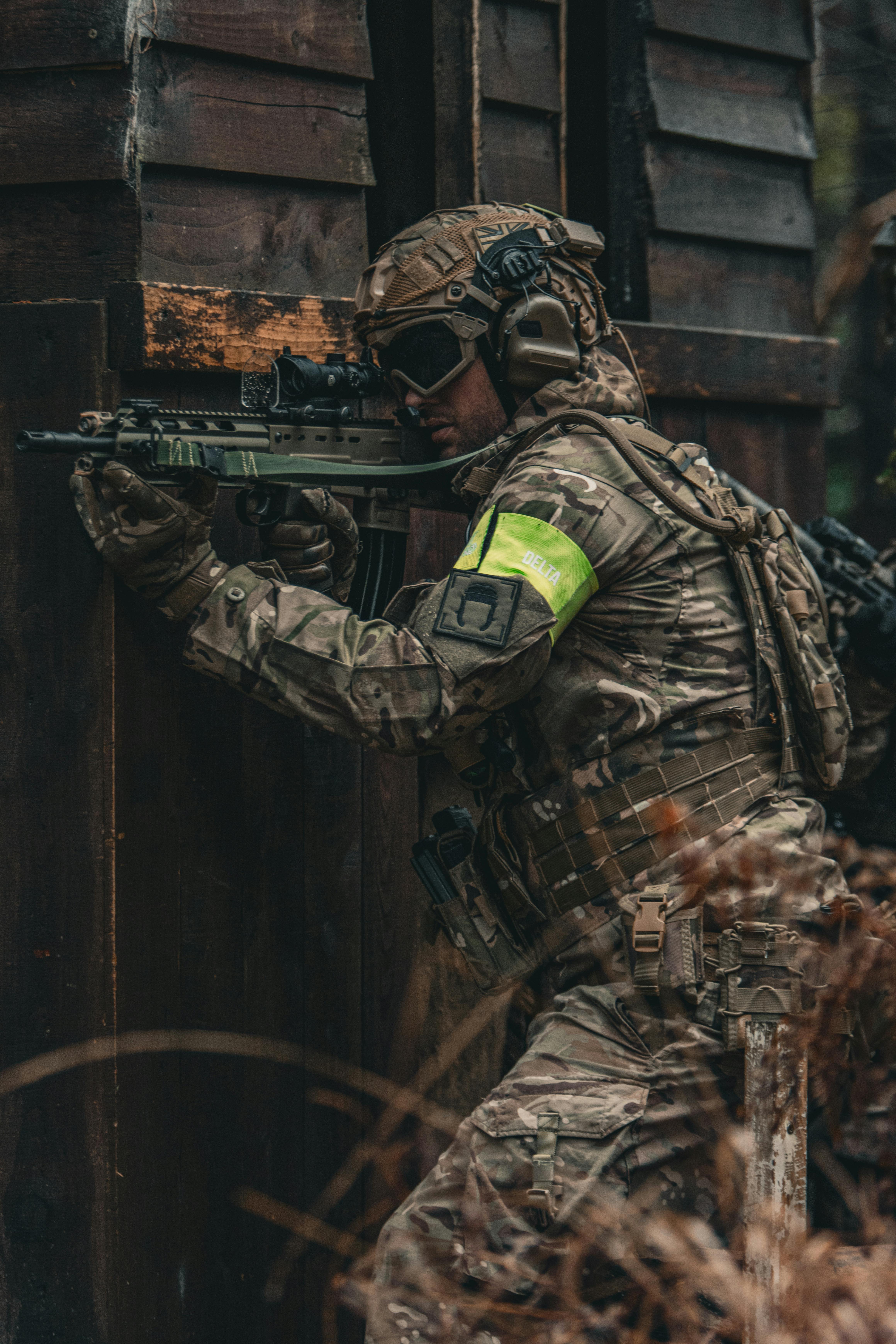 A soldier in full camouflage uniform aims a weapon outdoors, demonstrating military readiness.